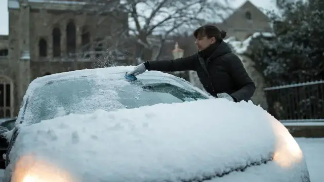 A woman wipes snow off the windshield of a car.