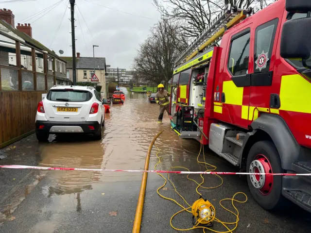 A fire engine parked in deep brown water with a fireman working at the back of the engine. They are in a a narrow residential street beside a parked white SUV and some terraced houses.