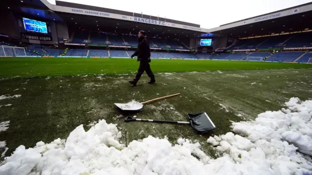 A member of grounds staff walks past a pile of snow. Two snow shovels have been laid on the ground.