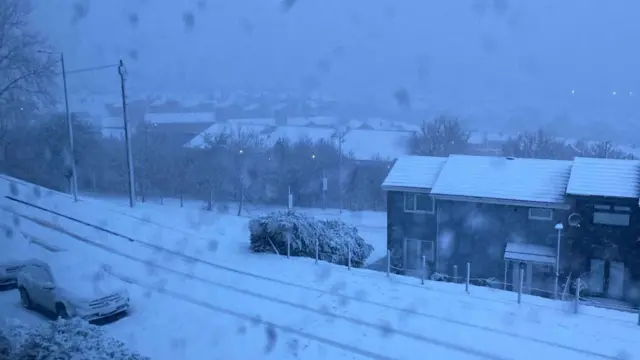 A road, cars and a row of terraced houses are covered in snow. Snow is falling