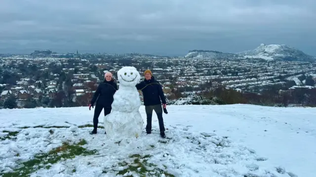 A man and a woman are smiling as they stand next to a snowman.