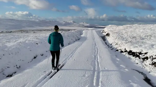 A person skis down a track. There is deep snow on the ground and snow-covered mountains in the distance.