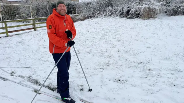 Gareth is wearing a hat and winter clothing. He is holding ski poles and skis on his feet.