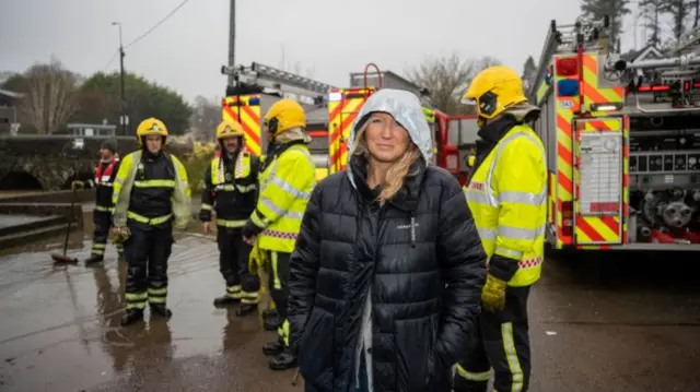 Rebecca Mullen, with a thick black coat on and grey hood, is stood in front of two fire engines and five emergency workers - one who is holding a broom