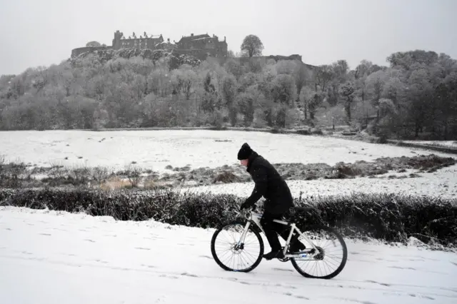 Cyclist in winter clothing and beanie riding past Stirling Castle in the background. Snow blankets the ground, trees and castle.