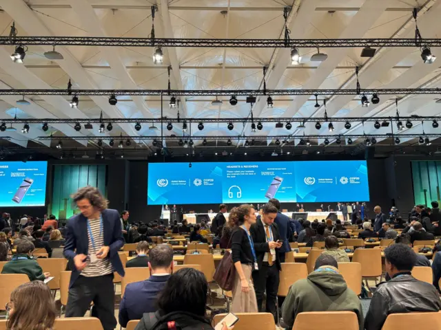 Stage of plenary with rows of wooden seats in front, filled with people and large blue screens