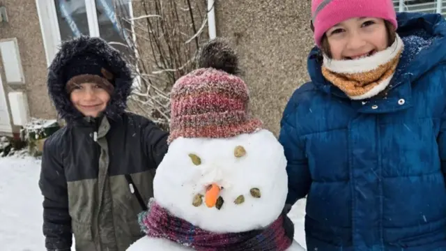 Two children smile next to the snowman they built