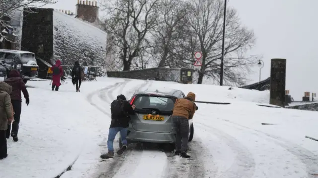 People help to push a vehicle uphill in the snow at Stirling Castle, Stirling, Scotland. One man (L) is in brown boots, jears and black winter jacket, the other (R) is in black jeans, light brown jacket and black boots