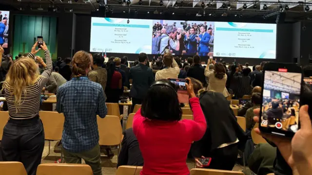 Room clapping at COP29 as people stand up, facing stage, clapping their hands and taking pictures on their phones