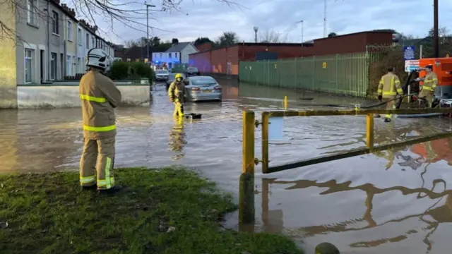 A flooded residential street near Belfast, where four firefighters are stood trying to pump water away from the streets