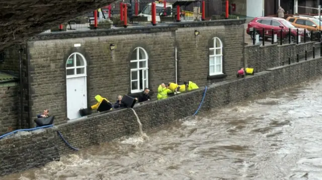 People remove water from street near the river in Pontypridd, South Wales  using plastic buckets. Most of them are wearing yellow hi-vi jackets and hats. Parked cars visible at top right of the frame