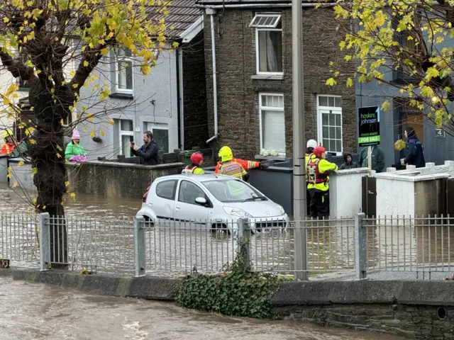 Car submerged in water as firefighters speak to residents outside a row of houses in Pontypridd