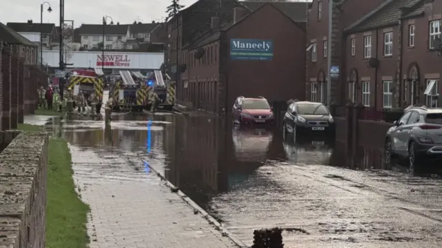 Flooded street in Northern Ireland after Storm Bert passage. Firefighters engaged in rescue operation can be seen in the background, their engines in high water. There's parked cars to the left of the frame also in high water