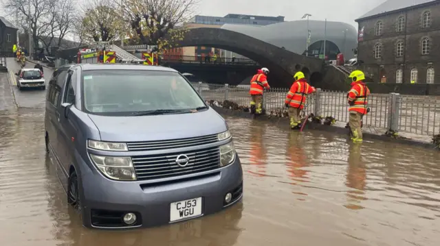 Firefighter pump water out of a street. There is a car at the front of the image that is stuck in floodwater