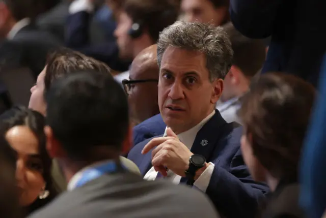 UK energy minister Ed Miliband at COP29 in Baku. He is seated and in focus, with the backs of other attendees out of focus. He's turning around and looking at the camera, from the chest up, wearing a dark suit