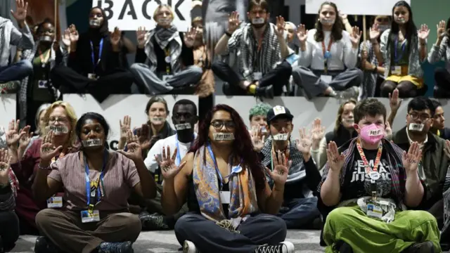 Activists sitting in rows with their hands up and tape on their mouths with the words "pay up".