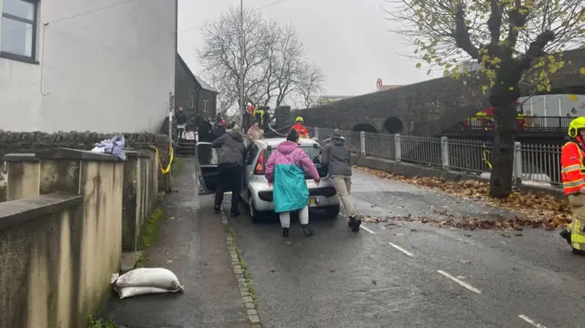 Residents on a street in Rhondda Cynon Taf are pushing a car. There are firemen on the road also.