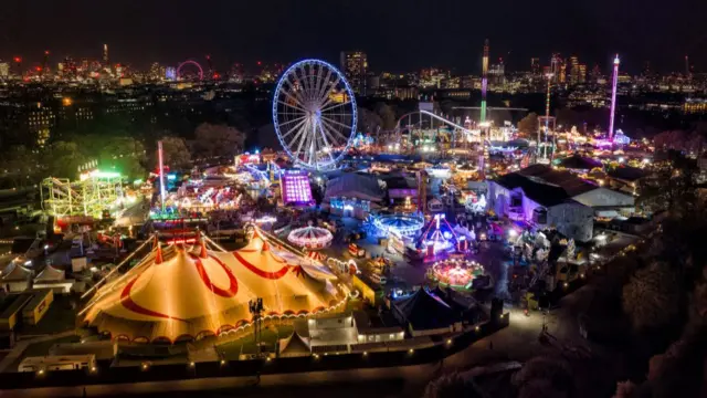 Winter Wonderland fair in Hyde Park, aerial view by night. A white circus tent with red decorations visible at bottom left of image. Several rides visible including a ferris wheel