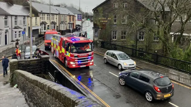 Fire engine on road in Pontypridd. Two cars (one grey and one black) also driving over what appears to be a stone bridge