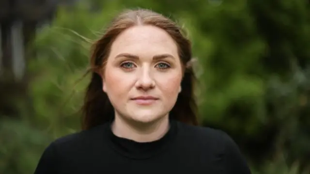 Laura Young, a climate activist and environmental scientist, shot from shoulders up, wearing a dark top, looking at camera, with blurred greenery in background