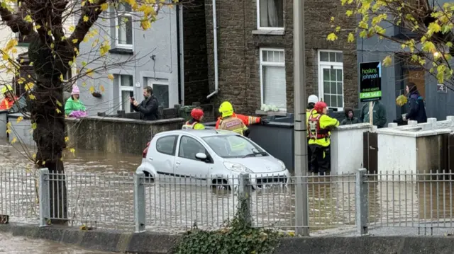 Firefighters in water rescue gear speak to residents of flooded street. A woman in rain jacket can be seen waiting outside a door with a man and a child