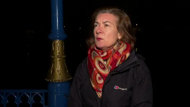 Welsh first minister Eluned Morgan in black coat and red scarf facing towards camera, standing next to blue pole and fence at night in darkness