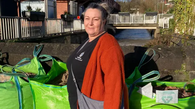 Tasha Saunders stands next to three large green sandbags. She is pictured in front of a river