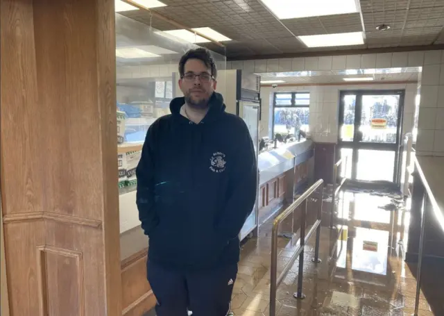 Ben Ruddy in navy tracksuit, looking at the camera. He is stood inside his fish and chip shop, which is flooded near the door