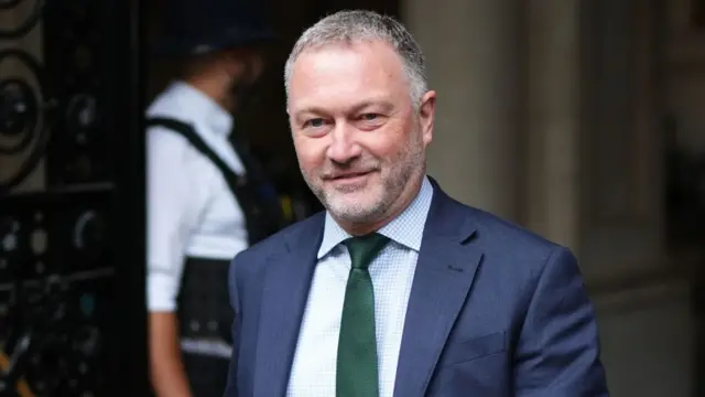 Steve Reed walking through a black gate. He is wearing a suit with a navy blazer and green tie. He had greying hair and is smiling.