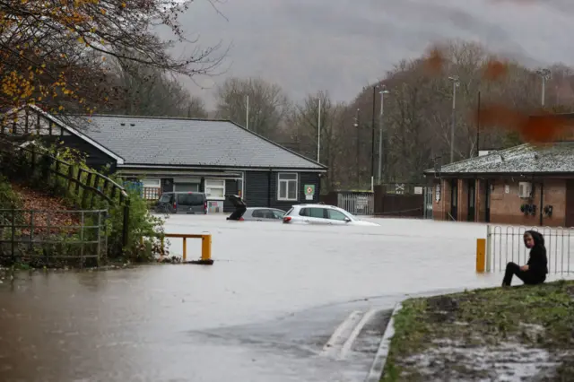 A person sits by the flooded water and there are submerged cars.