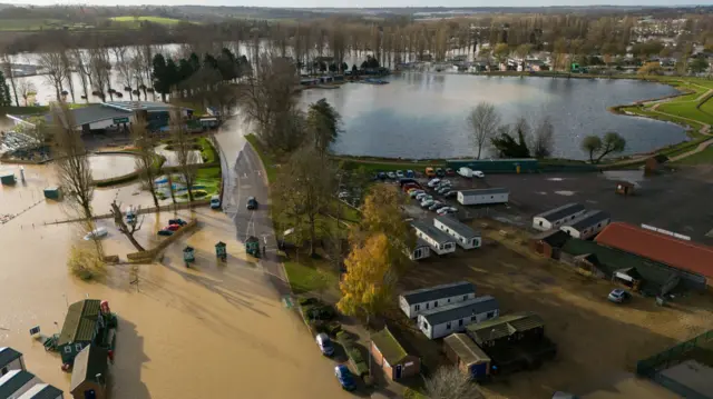 An aerial view of a flooded road and land, and also a lake. The lake has not flooded. Trees and mobile homes are visible near the flood water.