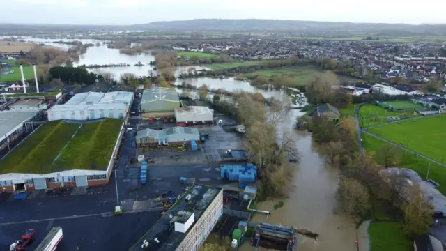 River with burst banks flowing between a sports pitch and a small industrial estate. The river stretches into the background and is flooded all the way to the back of the image