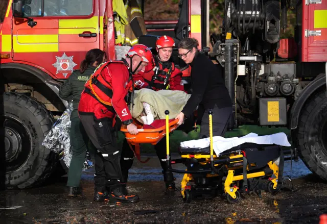 A local resident, wrapped in blankets, is attended to as she is lifted into an emergency vehicle at the Billing Aquadrome.