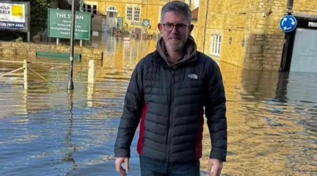 Man with glasses and a warm coat standing in front of a flooded hotel. The sign reads The Swan.