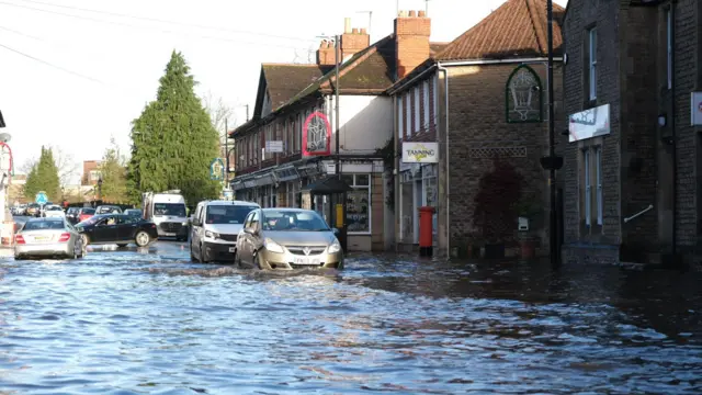 Cars driving through flood water, which rises about halfway up their wheels.