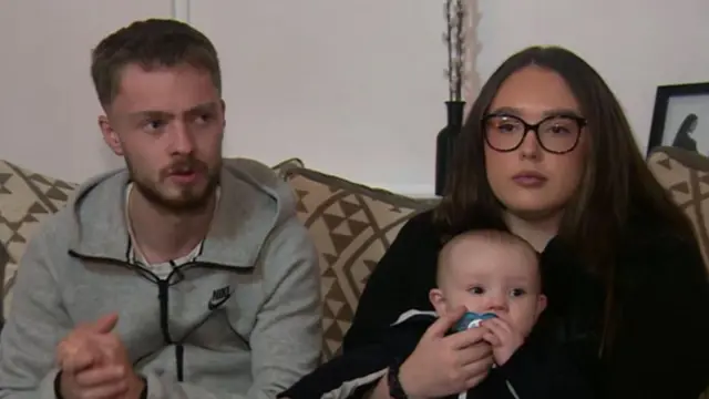 Andre Randles with partner Paige Newsome and their son Luca sat on a sofa. Randles is wearing a grey zip-up jumper and Newsome is wearing a black jumper with black-rimmed glasses. Luca has a blue dummer in his mouth and is sat on Newsome's lap.