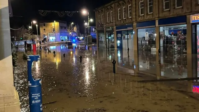 The high street in Chippenham underwater. The flood water can be seen covering the bottom of shop windows and doors. Bollards down the high street are also almost completely submerged, with just the tops showing.