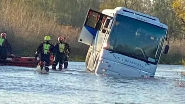 A bus is seen leaning to one side in flood water, with fire and rescue personnel stood alongside it with a small dinghy.