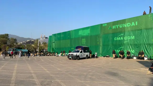 Soldiers with riot shields standing in front of a wall built of containers and some green tarp