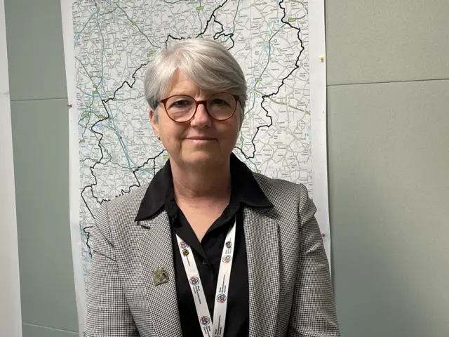 A bespectacled woman wears a white-black checkered jacket and black shirt standing in front of a map of Northamptonshire