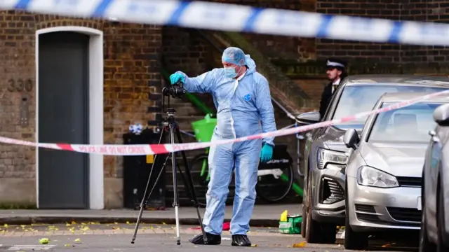 A police officer in forensic scrubs takes a photo with a camera on a tripod in the street