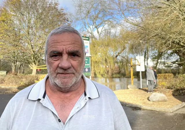 A man in a open grey polo shirt stands in front of a flooded road