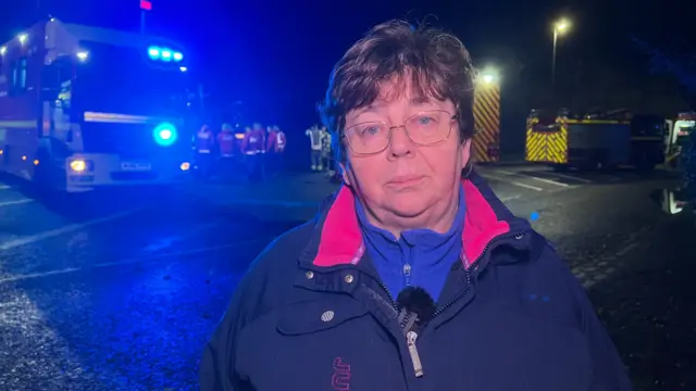 Councillor Cheryl Kirby stood on a road in front of a fire engine. It is dark and the blue emergency services lights can be seen in the background. Ms Kirby is wearing a thick blue waterproof coat.