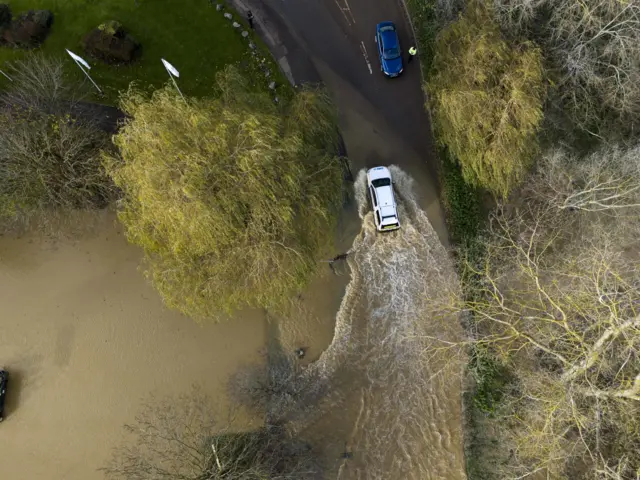 A drone shot of a white car driving through deep brown flood water. A blue car stops on the approach on the other side of the road