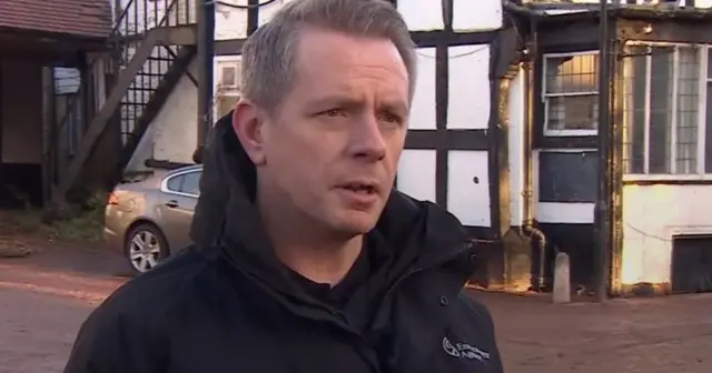 A man wearing a black jacket is stood in a town centre with a black and white building behind him.