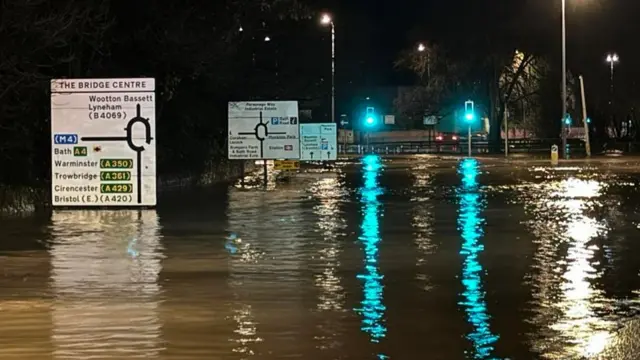 A road approaching the Bridge Centre Roundabout which is completely covered in flood water. It is dark and traffic light reflections can be seen in the water.