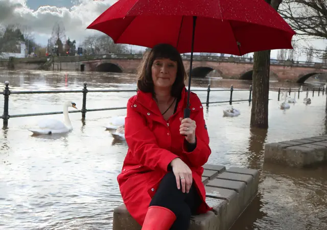 A woman in a red coat and holding a red umbrella sits by the River Severn.