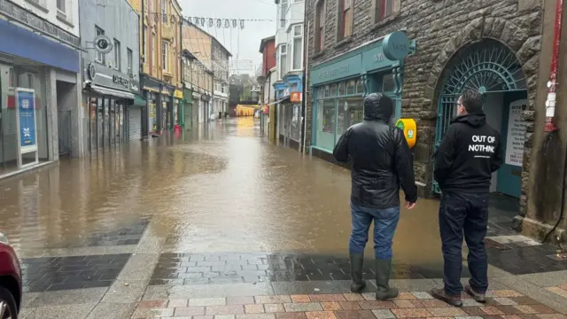 Floodwater runs down the middle of a street in Pontypridd as two men standing with their backs to the camera look on.