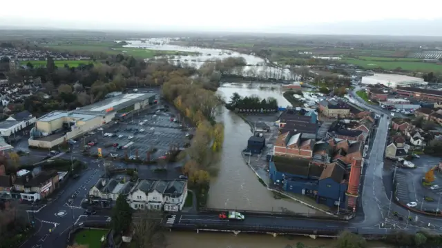 River with broken banks snaking its way between a car park and a series of low-rise buildings. A road bridge crosses it, with a van atop it.