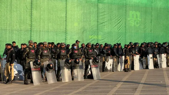 Soldiers with riot shields standing in front of a wall built of containers and some green tarp